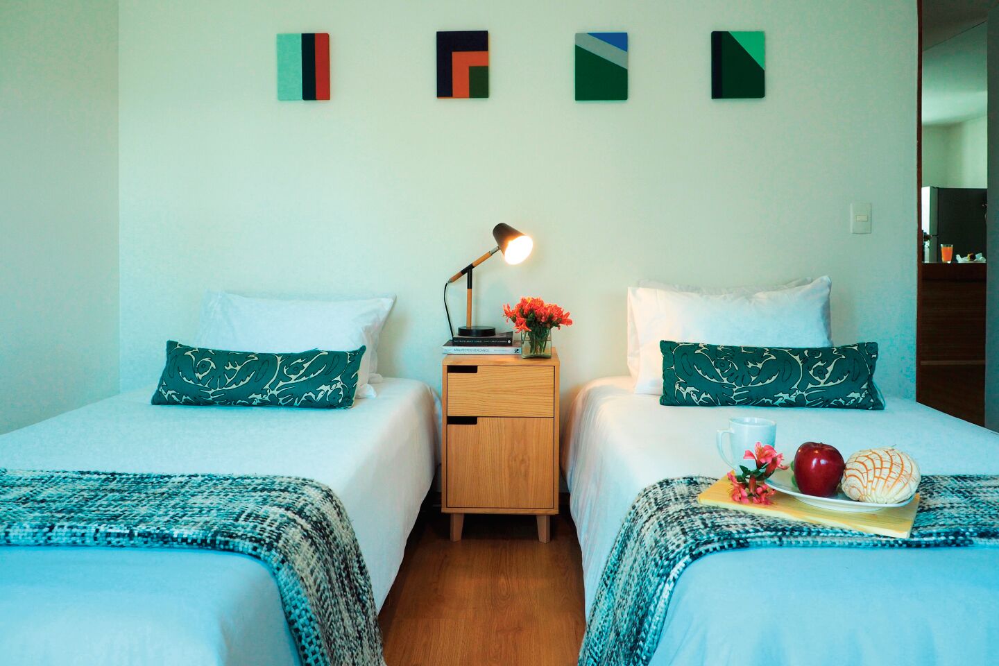 Una habitación de hotel con dos camas, cada una con un cojín estampado en verde, una mesita de noche con lámpara y una bandeja con frutas y flores.