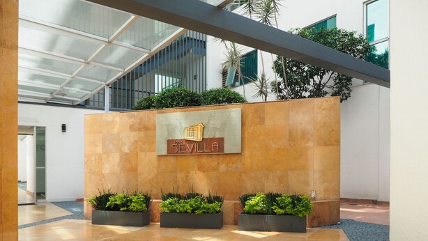 A modern lobby with a glass ceiling, a sign reading "SEVILLA," and three planters with greenery. - Torre Sevilla by The Local Way (Mexico City)
