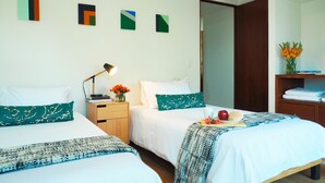 A hotel room with two beds, a nightstand with a lamp, a vase with flowers, and a shelf with folded towels. - Torre Sevilla by The Local Way (Mexico City)