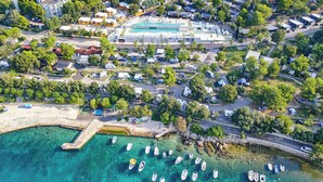 Vue aérienne d’une zone côtière comprenant un port de plaisance, plusieurs petits bateaux et divers bâtiments, dont un grand hôtel doté d’une piscine.