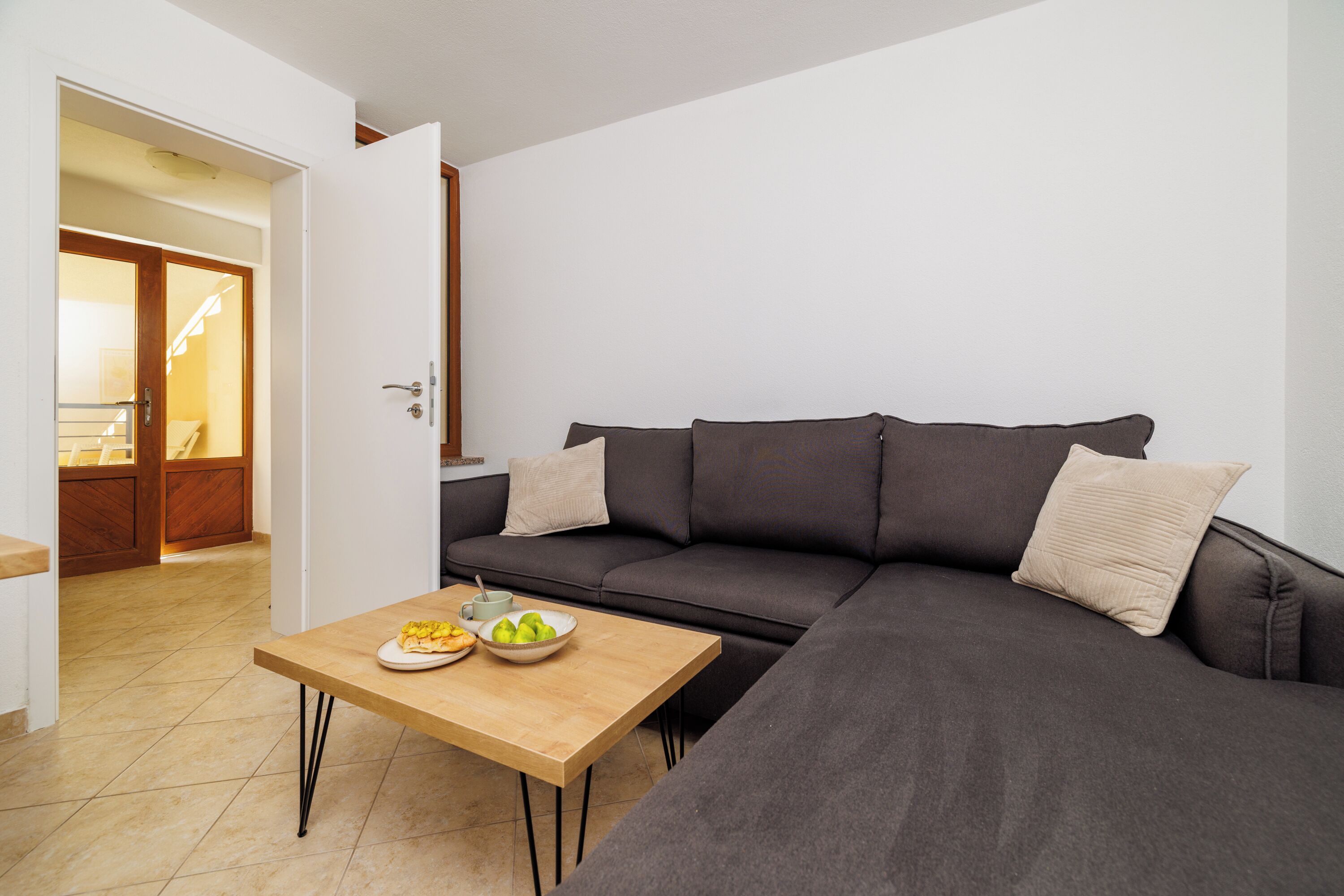 Una sala moderna con un sofá seccional gris oscuro, una mesa de centro de madera con un cuenco de fruta, y una puerta blanca que da a una cocina.