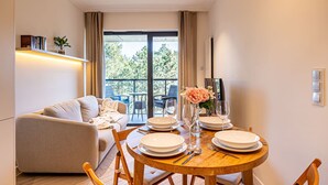 Ein Essbereich mit einem runden Holztisch, gedeckt für vier Personen, einer Couch und einem Balkon mit Blick auf Grünflächen.