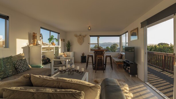 A spacious living room with a sofa, a coffee table, a TV, and a dining area with stools. There is a large window offering a view of the outdoors.