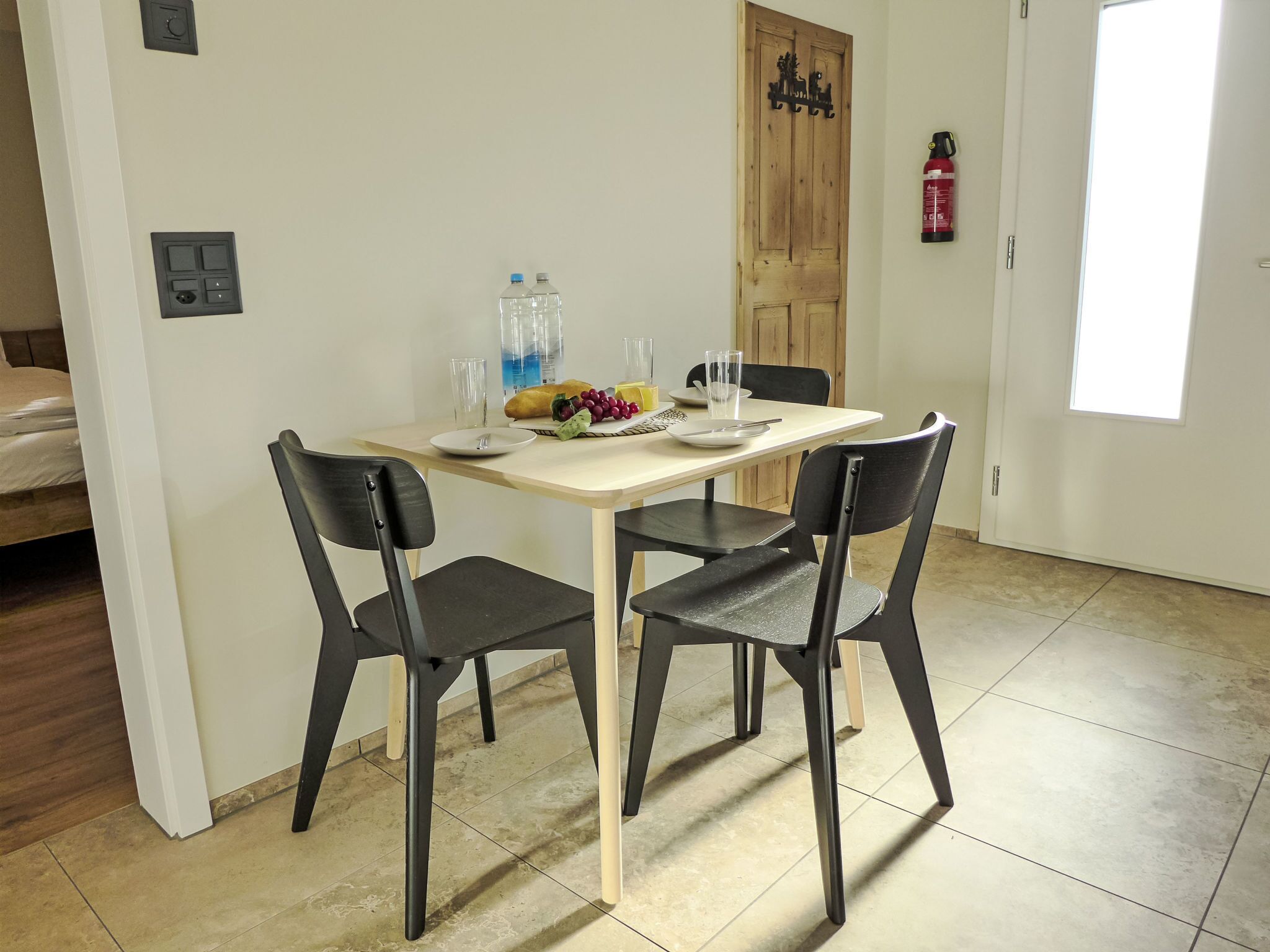 A dining area with a table set for two, two chairs, a fruit bowl, water bottles, and a fire extinguisher on the wall.