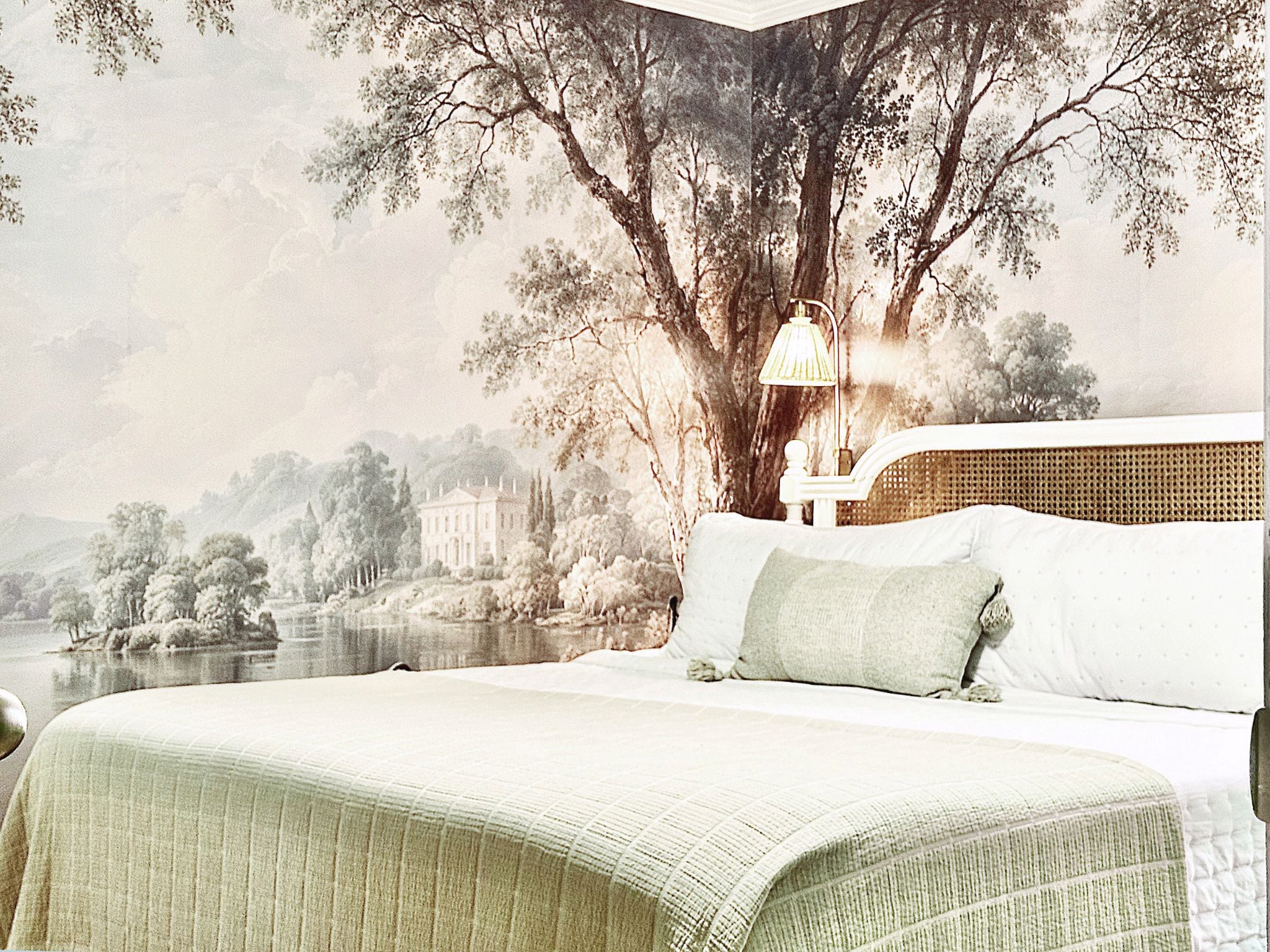 Une chambre à coucher avec un grand lit, une table de chevet, une lampe et une fresque murale représentant un paysage.