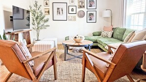A living room with a green sofa, two wooden chairs, a coffee table, a TV, and framed pictures on the wall. - Curated & Caffeinated! Gem near Duke & Downtown • Espresso bar, Eco+, Fire Pit (Durham)