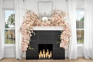 A fireplace with a floral arrangement above it, a mirror above the fireplace, and a view of a house through the window to the left. - 2 Linked Suites • 6BR/6BA • Heated Pool/Spa • BBQ (Houston)