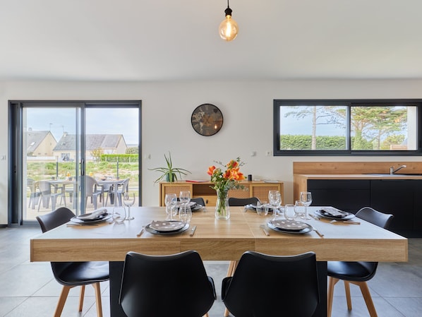 A modern dining area with a large wooden table set for four, surrounded by black chairs. A vase with orange flowers is on the table, and a world clock is on the wall.