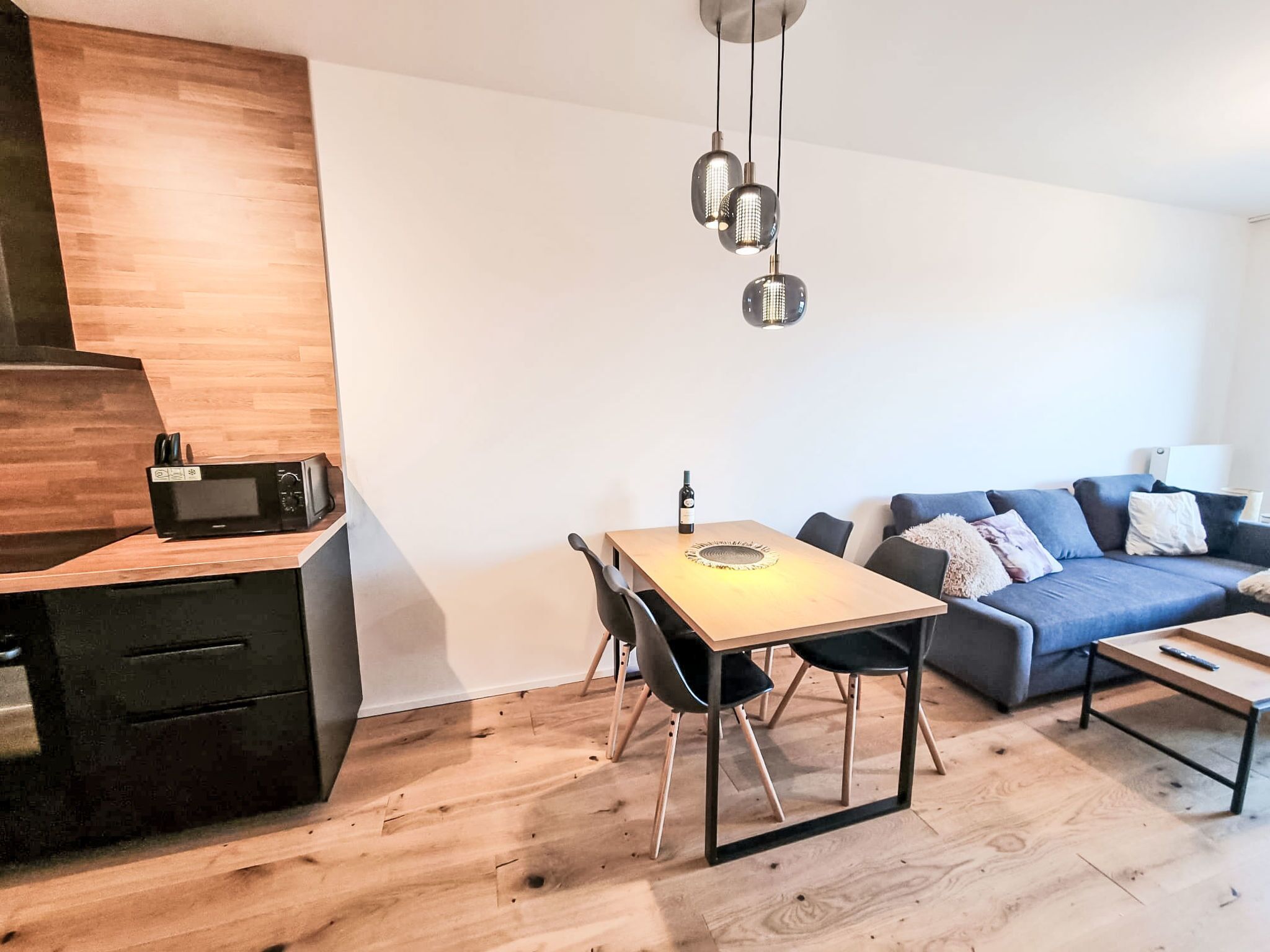 Une cuisine moderne avec un plan de travail en bois, un four à micro-ondes encastré et une machine à café. On y trouve une table à manger avec des chaises, ainsi qu’un canapé bleu dans l’espace salon.