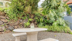 Un banco y una mesa de piedra en un jardín con una pared de piedra y diversas plantas.