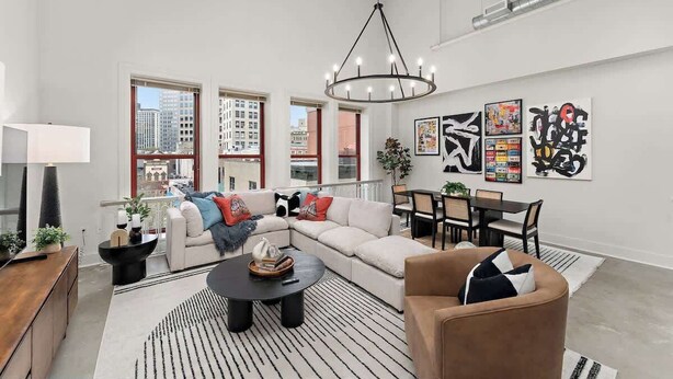 A modern living room with a sectional sofa, a round coffee table, and a dining area with a table and chairs. There are large windows providing a view of the cityscape. - Designer Loft Penthouse in the Heart of Downtown (Pittsburgh)