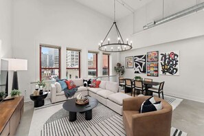 A modern living room with a sectional sofa, a round coffee table, and a dining area with a table and chairs. There are large windows providing a view of the cityscape. - Designer Loft Penthouse in the Heart of Downtown (Pittsburgh)
