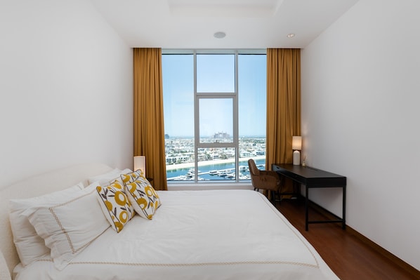 A hotel room with a large bed, a desk, and a view of a cityscape through floor-to-ceiling windows.