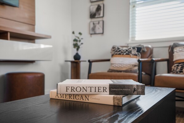 Une pile de livres intitulés « FRONTIER » et « AMERICAN » est posée sur une table en bois dans un salon, où se trouvent des fauteuils en cuir et une fenêtre avec des stores.