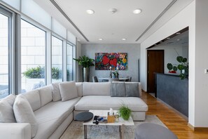 A modern living room with a large sectional sofa, a coffee table, and a dining area with a table and chairs. There are plants and a colorful abstract painting on the wall.