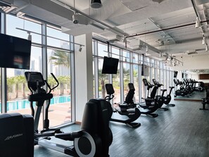 A gym with treadmills, stationary bikes, and exercise equipment. Large windows offer views of palm trees and a pool. - Cozy DIFC Duplex w/ Burj View, Pool and Gym (Dubai)
