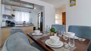 A modern dining area with a wooden table set for four, a grey sofa, and a kitchen in the background. - Spacious 3-bedroom apartment with WiFi, swimming pool in vibrant Tatu City (Tatu City)