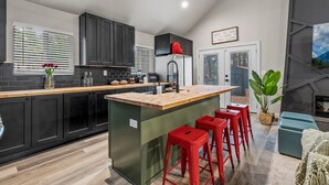 A modern kitchen with dark cabinets, a wooden island with stools, a large window with blinds, and a fireplace. - Pine Del | 3BR/2BA | Spend Spring in Flagstaff! (Flagstaff)