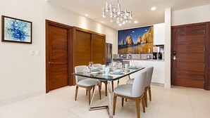 A dining area with a glass table, four chairs, and a chandelier. There are wooden doors and a kitchen in the background. - One Bedroom Suite Pool View, Los Cabos, Villa la Valencia Resort (San José del Cabo)