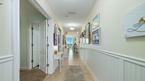 A hallway with white walls, beige tiles, and a beige carpet. There are framed pictures on the walls and a chandelier on the ceiling. - ESCAPE TO THE INDIES CONDO IN FORT MORGAN ALABAMA! (Gulf Shores)