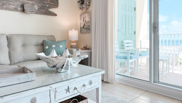 A living room with a glass coffee table, a sofa, and a view of a balcony with outdoor furniture. - ESCAPE TO THE INDIES CONDO IN FORT MORGAN ALABAMA! (Gulf Shores)