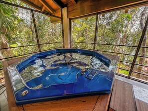 Un bain à remous avec une terrasse en bois et une vue sur une forêt, visible à travers de grandes fenêtres.