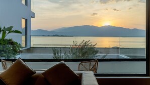 Une chambre moderne dotée d’une grande fenêtre donnant sur un plan d’eau et des montagnes, avec une vue particulièrement belle au coucher du soleil.