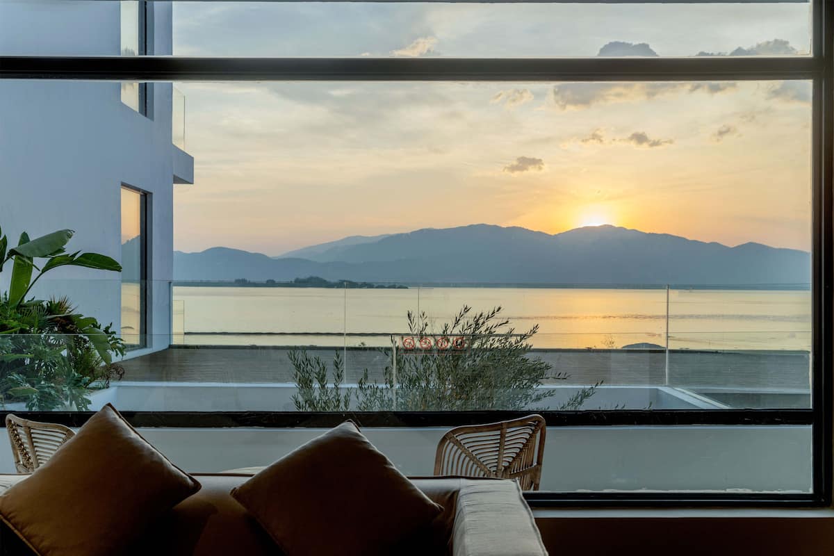 Une chambre moderne dotée d’une grande fenêtre donnant sur un plan d’eau et des montagnes, avec une vue particulièrement belle au coucher du soleil.