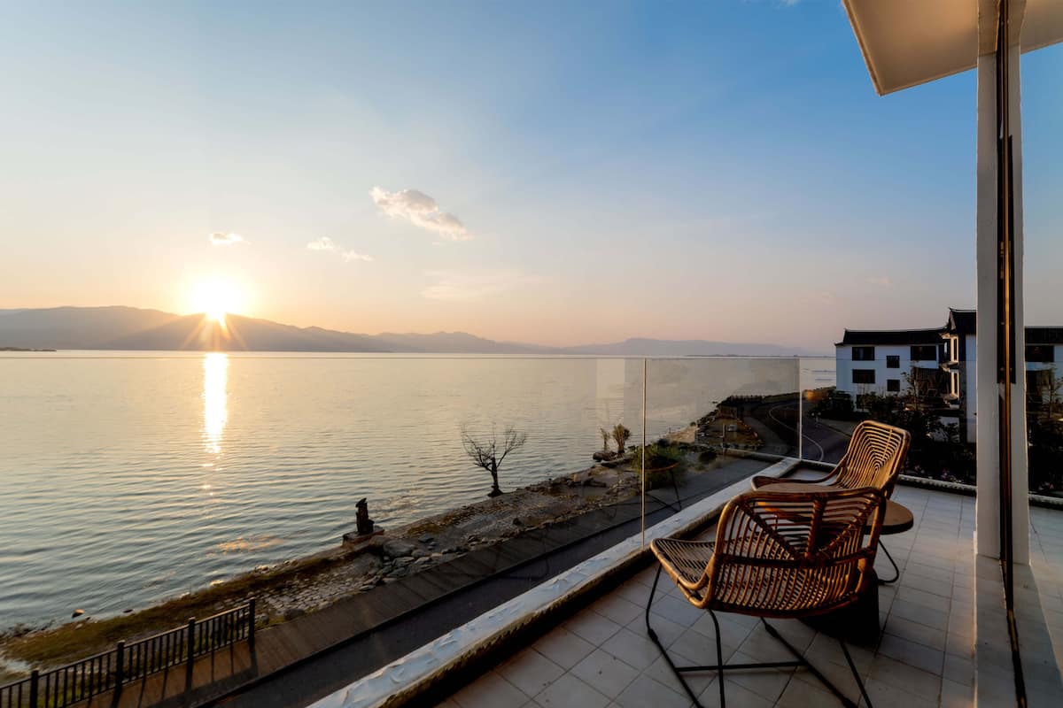 Un balcon donnant sur un lac paisible au coucher du soleil, avec des chaises en bois et une balustrade.