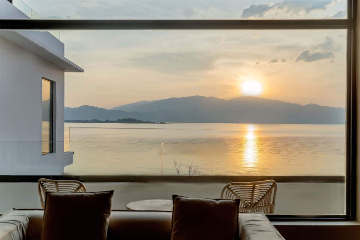 Une chambre moderne dotée d’une grande fenêtre donnant sur un lac paisible et des montagnes lointaines, le tout baigné par les couleurs du coucher de soleil.