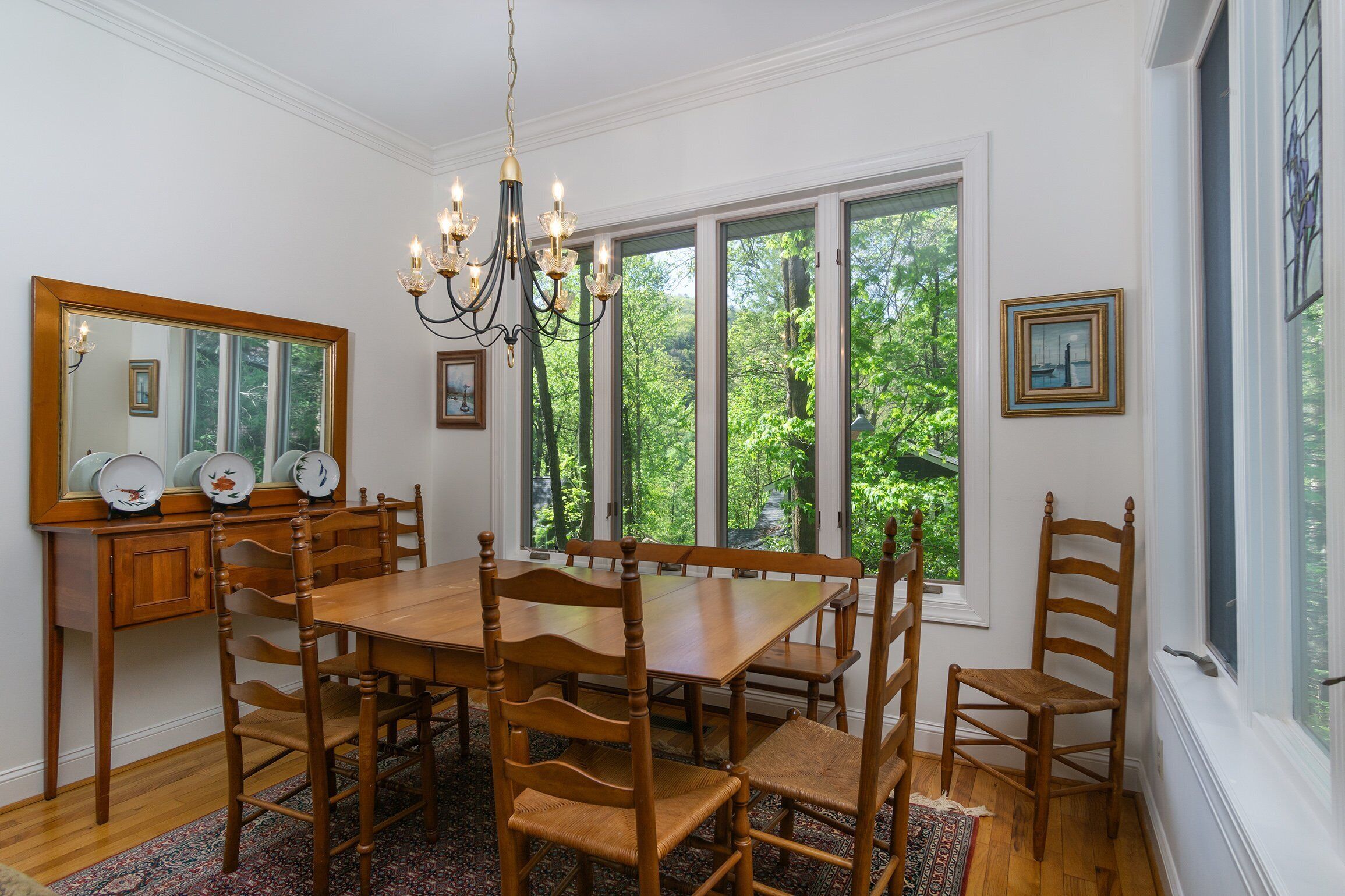 Un comedor con una mesa y sillas de madera, una araña, un espejo y un ventanal con vistas a árboles.