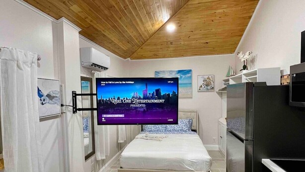 Un dormitorio con una cama, un televisor instalado en la pared, un refrigerador y techo de madera.