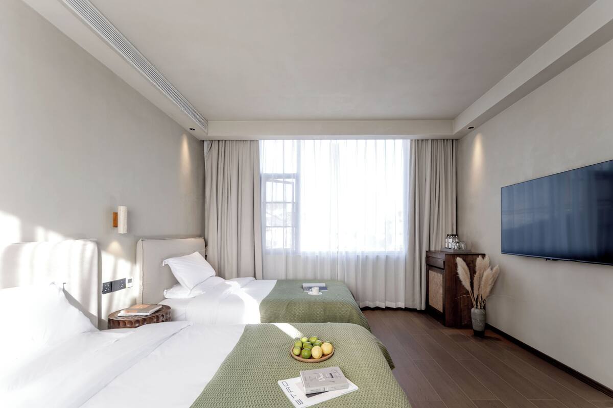 A hotel room with two beds, a flat-screen TV, a wooden side table, and a window with curtains.