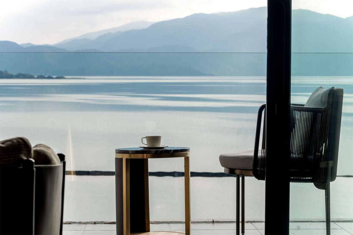 A room with a view of a lake and mountains, a table with a cup, and a chair.