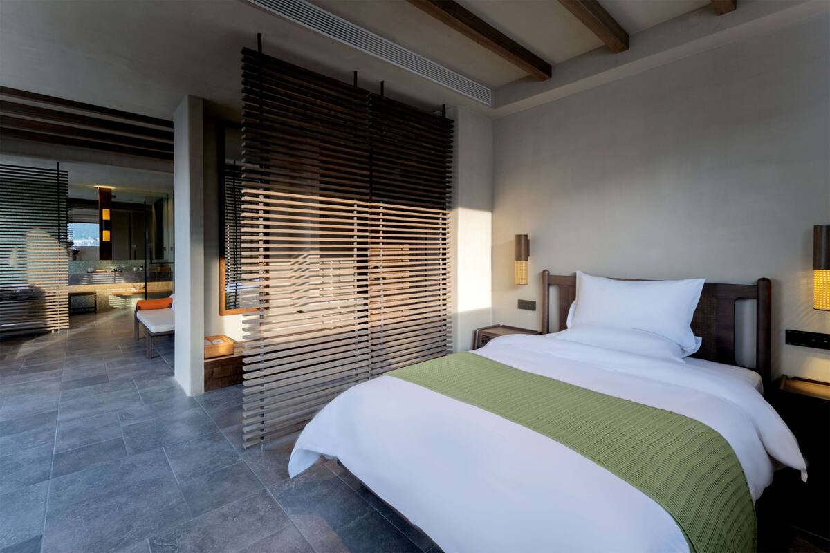 A modern hotel room with a large bed, wooden blinds, a green throw, and a view of the city.