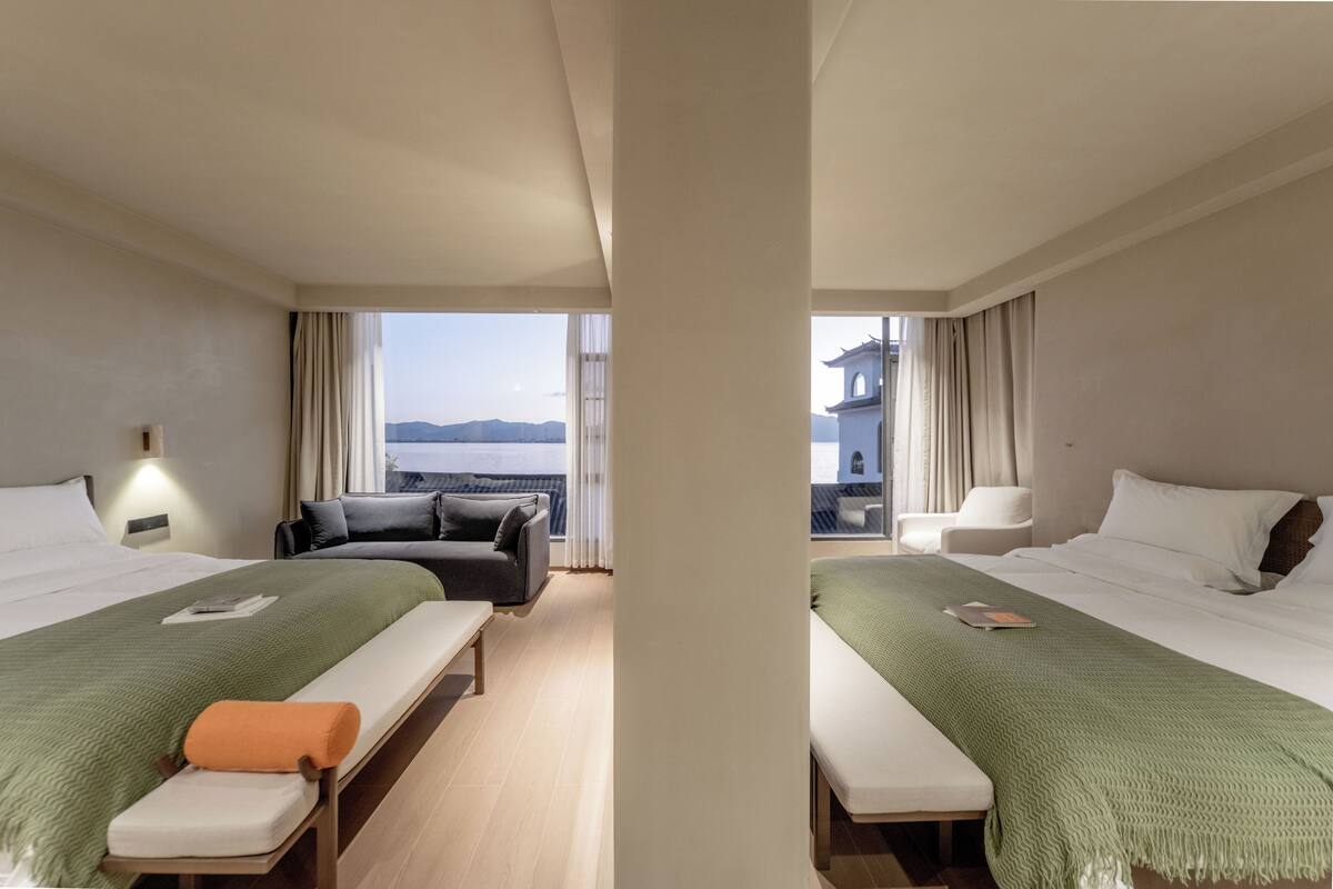 A hotel room with two beds, a sofa, a bench, and a view of the sea.