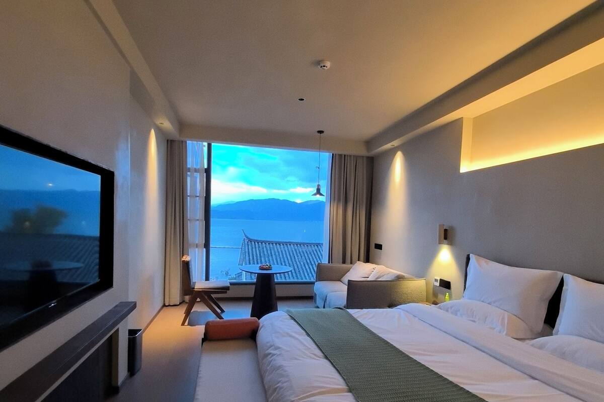 A modern hotel room with a large bed, a TV, a balcony with a view of the sea, and a small table.