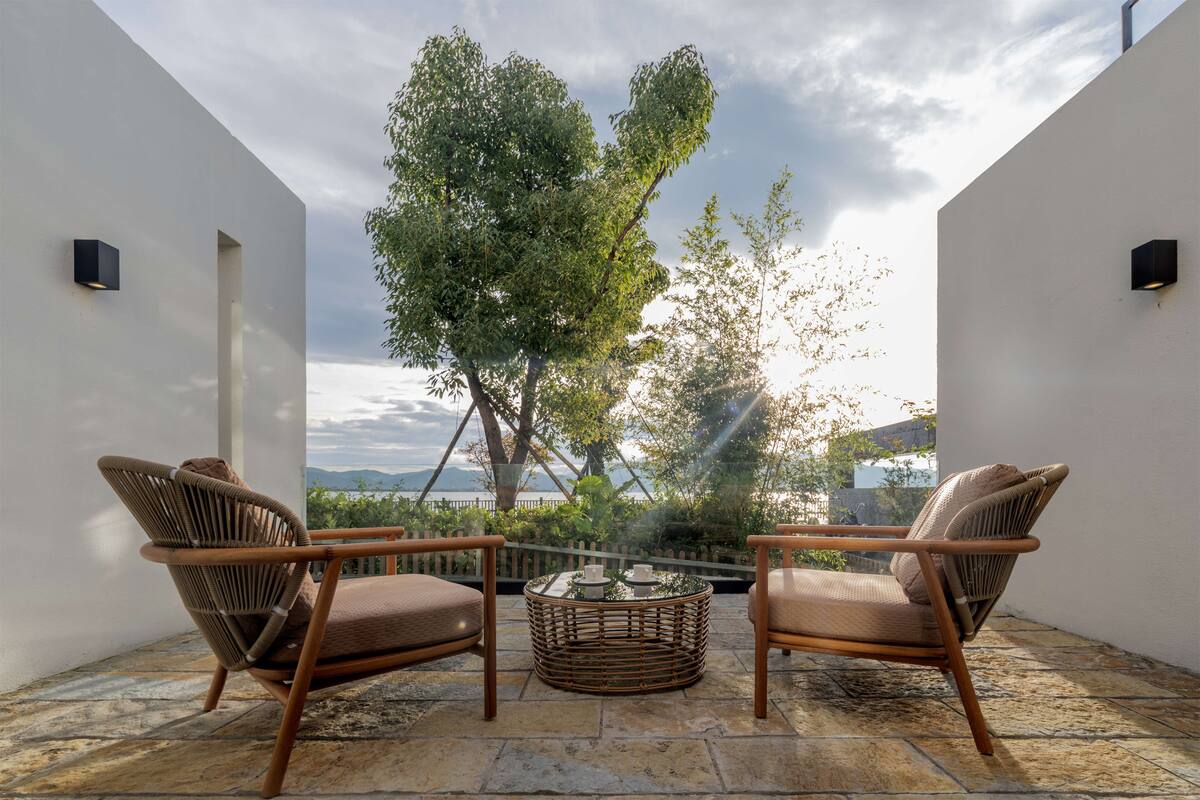 A patio with two wicker chairs and a round table, overlooking a tree and a view of the landscape.