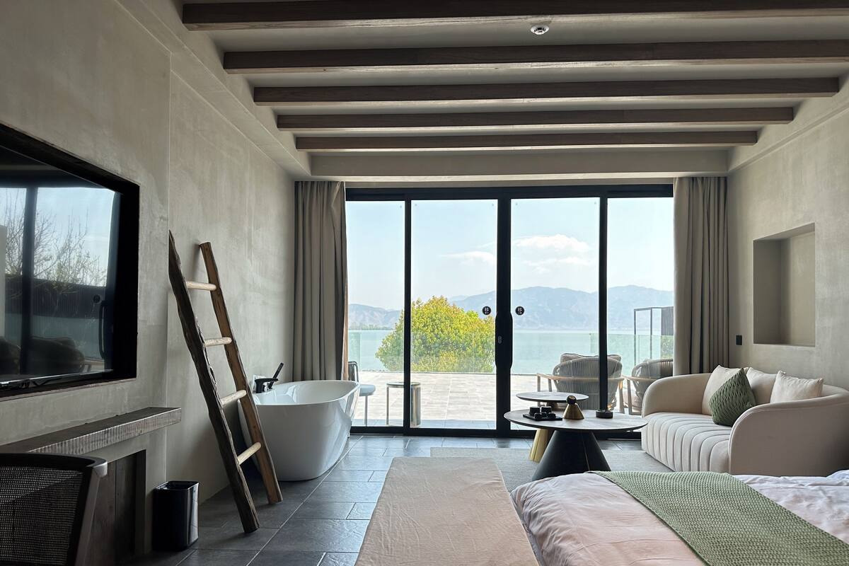 A modern hotel room with a large bed, a seating area with a table, a bathtub, and a balcony with a scenic view.
