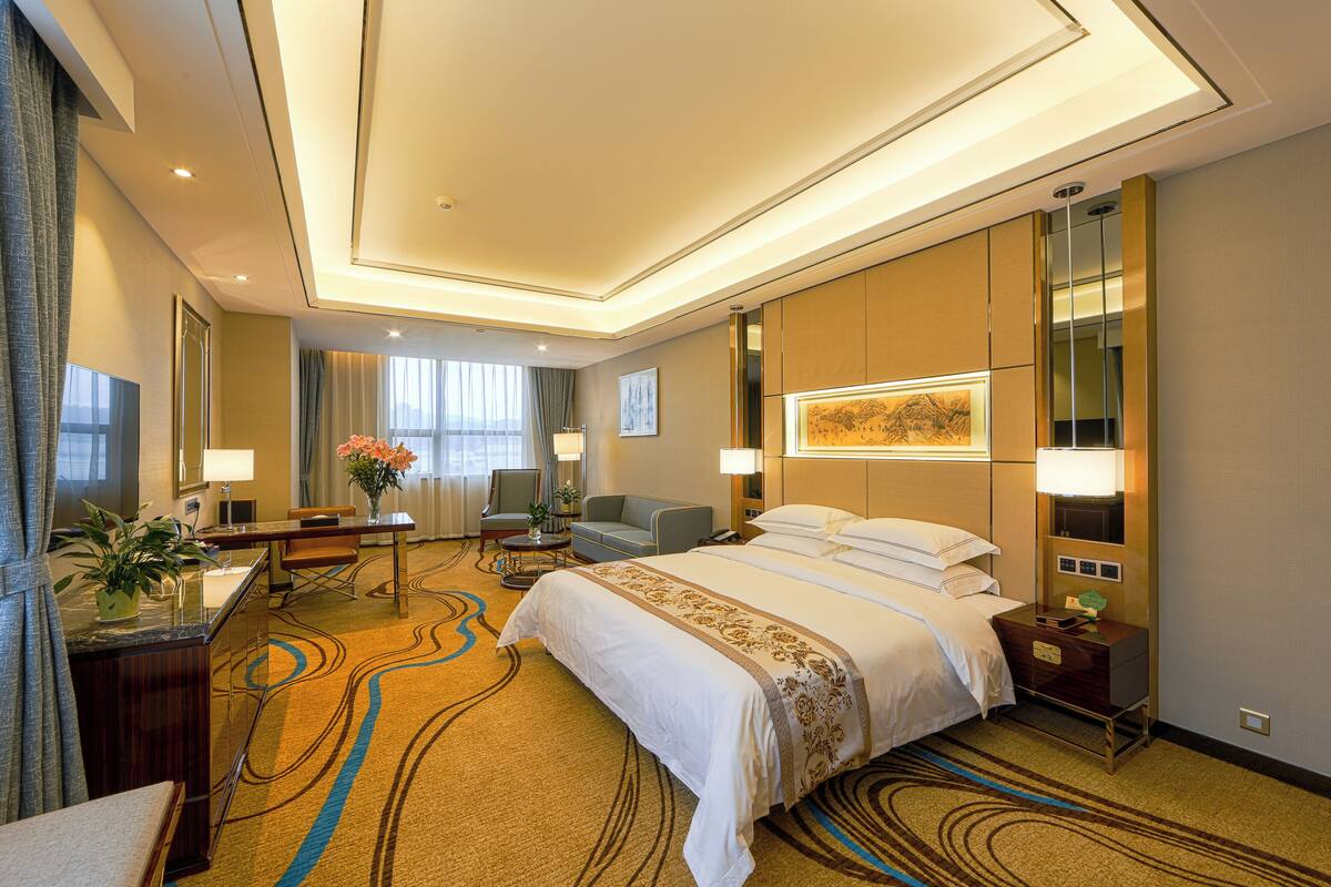 A hotel room with a large bed, a desk with a chair, a small table with a vase, a sofa, and a nightstand with a lamp.