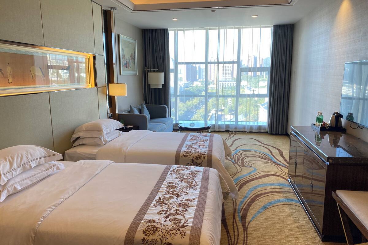 A hotel room with two beds, a desk, a chair, a lamp, and a large window with a city view.