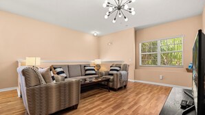 A living room with a sectional sofa, two armchairs, a coffee table, a TV stand with a flat-screen TV, a remote, and a ceiling light fixture.