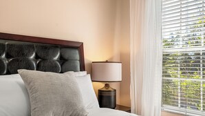A neatly made bed with white linens and a grey pillow, a dark headboard, a bedside lamp, and a window with blinds.