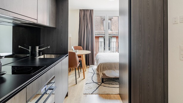 A modern kitchen with dark wood cabinets, a black countertop, and stainless steel appliances. There is a dining area with a round table and chairs, and a bed with a white duvet in the background.