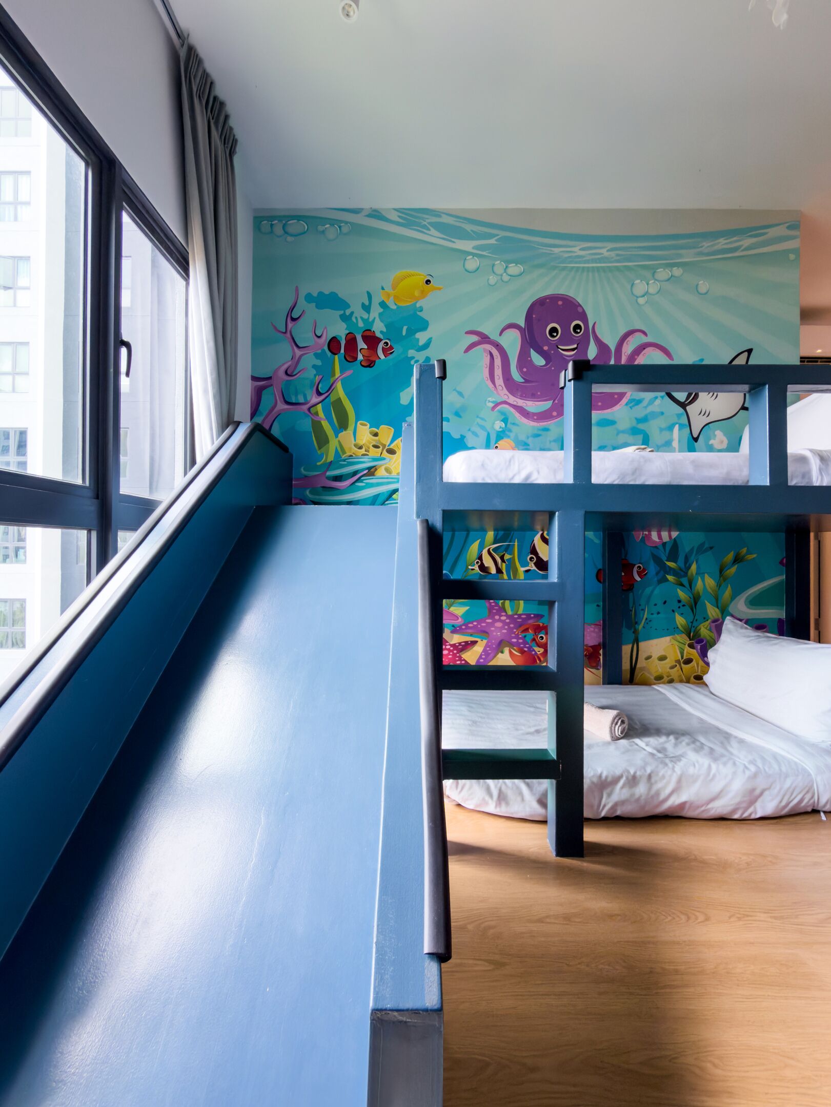 Una cama litera con tobogán, un ventanal grande y un mural de una escena submarina.