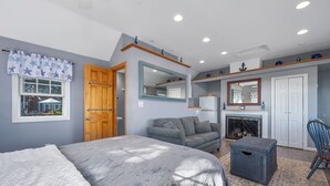 A bedroom with a bed, a gray sofa, a wicker chair, a fireplace, and a mirror.