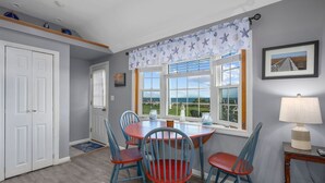 A dining area with a round table and four chairs, a window with a view of the sea, and a lamp on a side table.