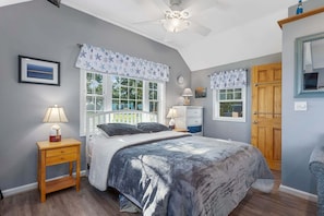 Une chambre à coucher comprenant un lit, des tables de chevet, une commode, un ventilateur de plafond et une grande fenêtre.
