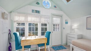 A room with a wooden table, two teal chairs, a bed, a window with lace curtains, and a ceiling with a skylight. - SeaGill Rest & Nest - A Peaceful Ocean Hideaway (South Yarmouth)