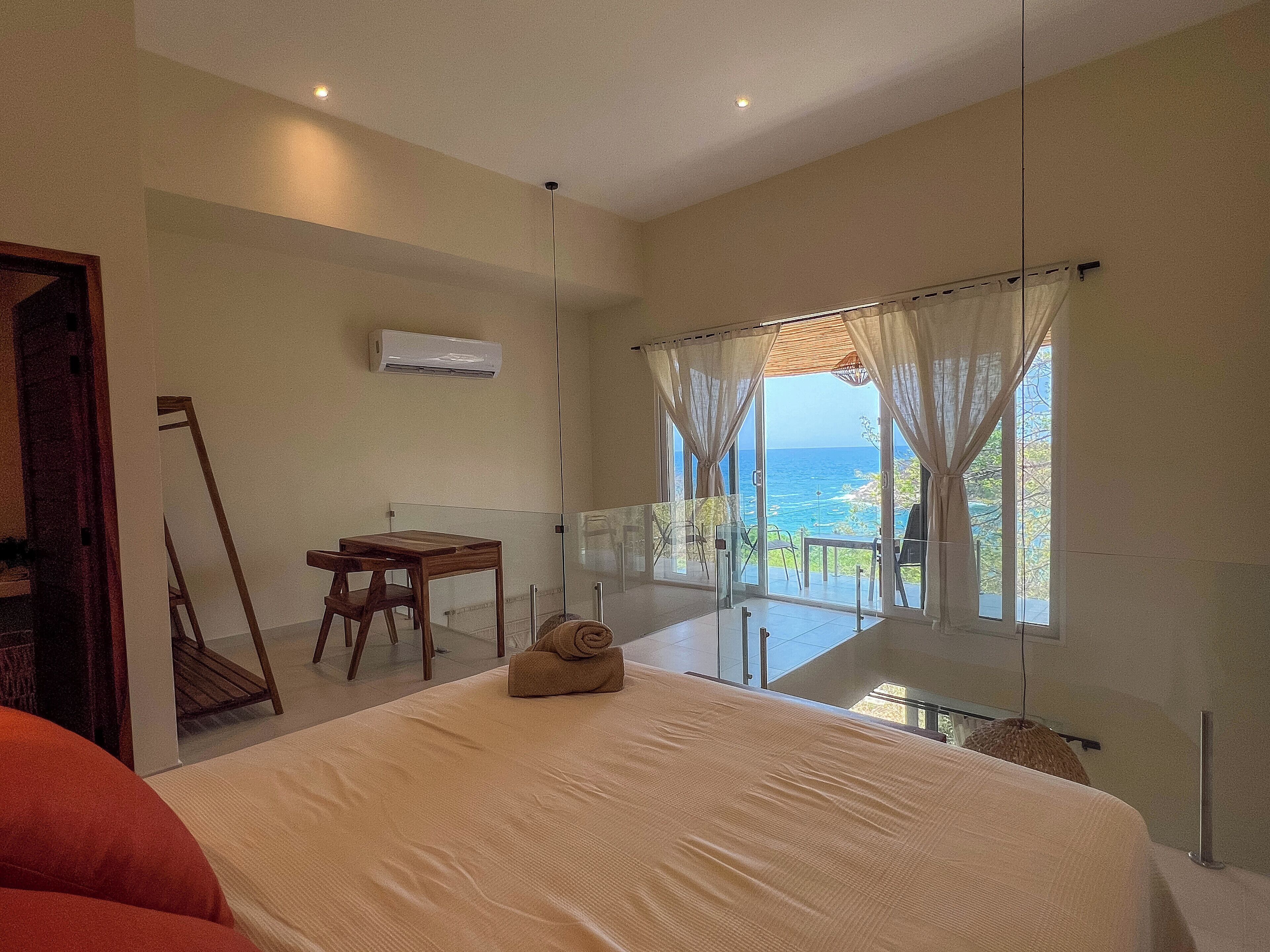 Un dormitorio con una cama grande, una silla de madera, una mesita y vistas al océano a través de amplias ventanas.